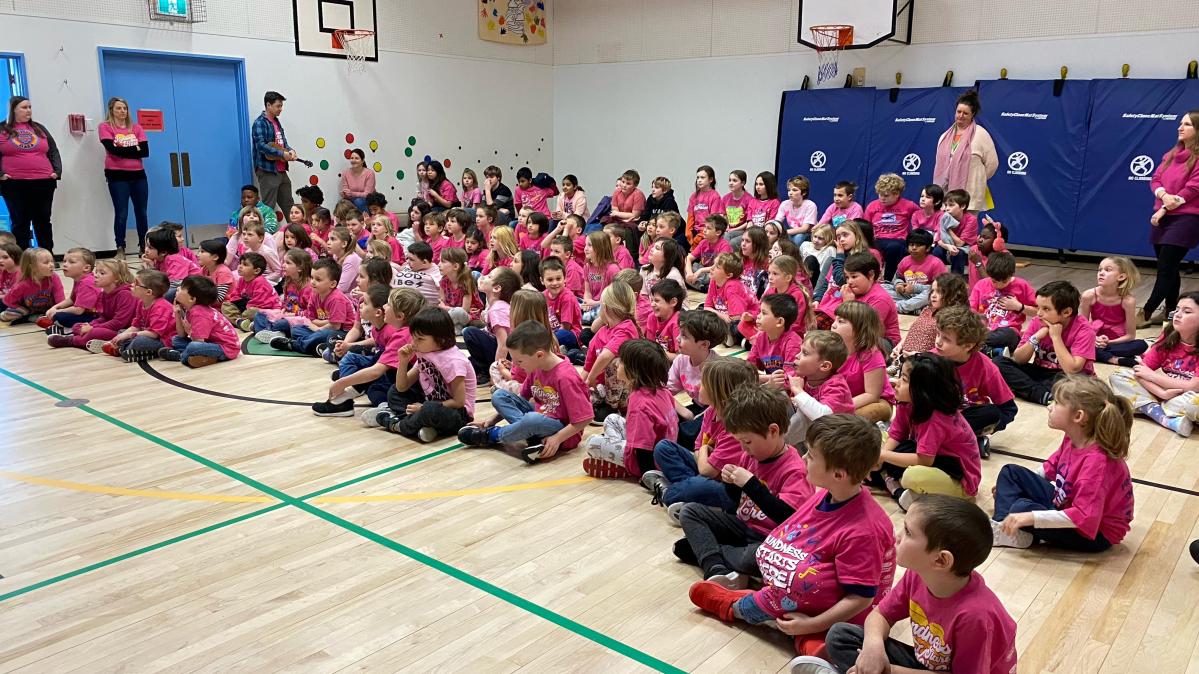 An elementary school class seated on a gymnasium floor, all wearing pink shirts.