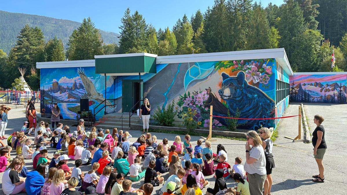 Rosemont's new mural! A group of elementary school students sit on the ground in front of a colourful school while a principal speaks to them.