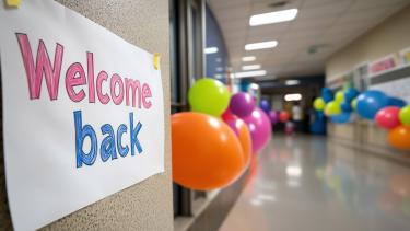 A handwritten "Welcome Back" poster on a hallway wall, with colorful balloons. The festive, celebratory of a school event or the first day back, with a blurred background of a school 