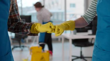 Custodians wearing yellow rubber gloves and blue aprons bump fists in an office setting while a custodian mops in the background